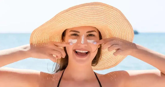Smiling woman applying best Korean face sunscreens while wearing a sun hat at the beach.