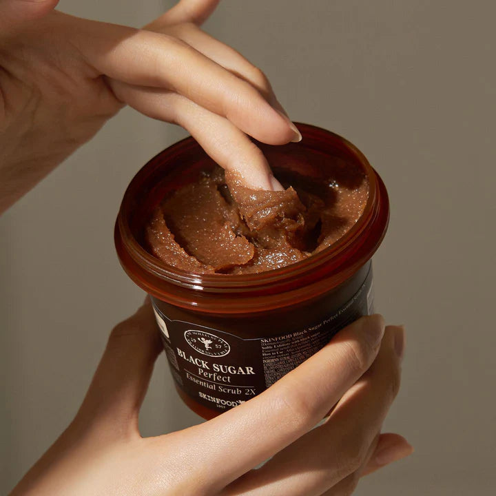 Hands holding a jar of SKINFOOD Black Sugar Perfect Essential Scrub 2X, showcasing its rich texture.