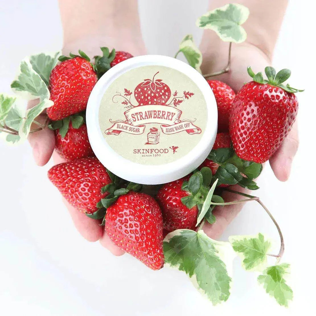 SKINFOOD Black Sugar Strawberry Mask Wash Off 100g surrounded by fresh strawberries and green leaves in hands.
